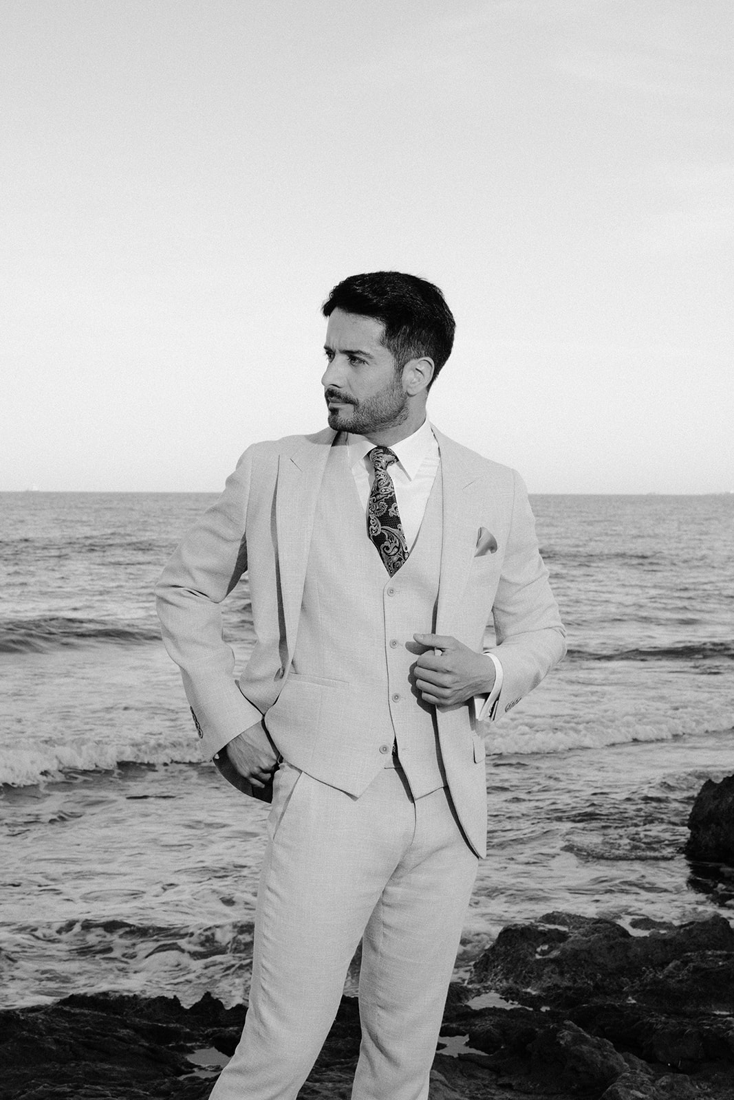 Handsome groom posing on the rocky shore in a chic wedding outfit, part of a Mediterranean love story shoot.